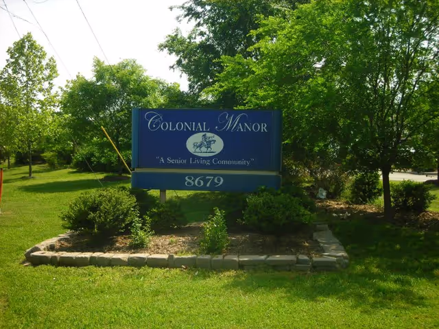 A blue Colonial Manor sign reading "A Senior Living Community" with the number 8679 set in a landscaped grassy area surrounded by trees.