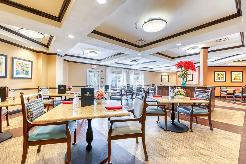 Bright and spacious dining room with multiple tables and chairs arranged neatly. Each table has a small flower arrangement and a menu labeled 'Pacifica'. The room features warm beige walls, framed artwork, large windows with white curtains, and modern ceiling lights.