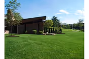 A well-maintained green lawn with a modern brick building on the left side under a blue sky with scattered clouds. There are trees and shrubs near the building and a few more trees in the background.