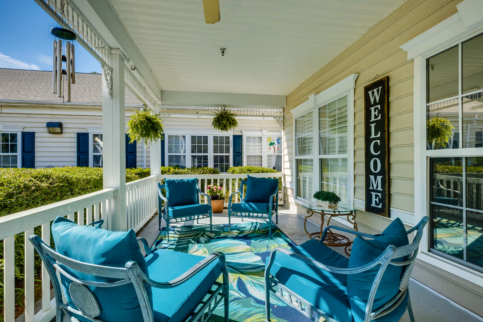 A covered outdoor porch area with teal cushioned chairs arranged around a glass-top table on a colorful leaf-patterned rug. Hanging plants and a wind chime decorate the porch, and a vertical wooden sign on the wall reads 'WELCOME'. The porch overlooks a garden with hedges and neighboring buildings with white siding and blue shutters.