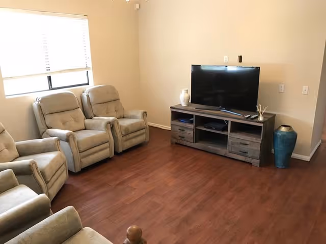 A living room with four beige recliner chairs arranged along the left wall near a window with blinds. Across from the chairs is a wooden TV stand holding a flat-screen television, a white vase, and some small decorative items. The floor is a dark wood laminate and the walls are painted a light beige color.