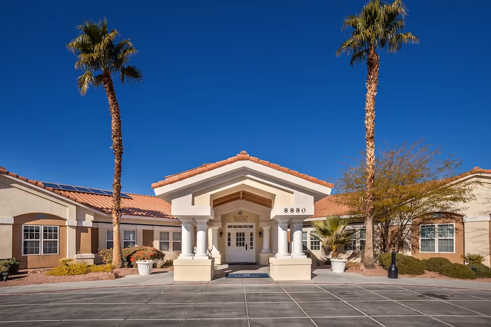 Front exterior view of a single-story building with a tiled roof and white columns at the entrance. Two tall palm trees flank the entrance, and there are shrubs and potted plants around the building. The sky is clear and blue.