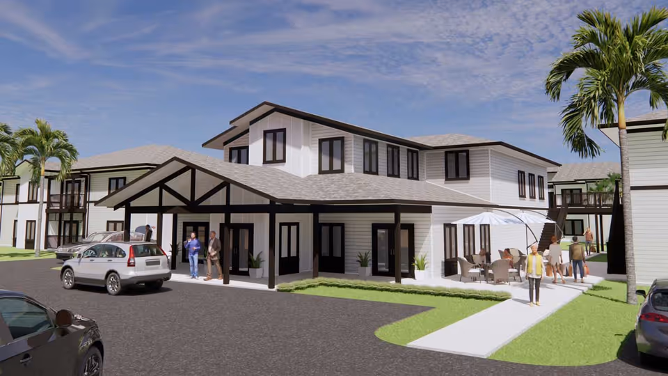 Exterior view of a two-story senior living facility with white siding and dark trim. Several people are seen walking and sitting outside near a covered entrance and patio area with outdoor furniture and an umbrella. Palm trees and parked cars are visible around the building under a partly cloudy sky.