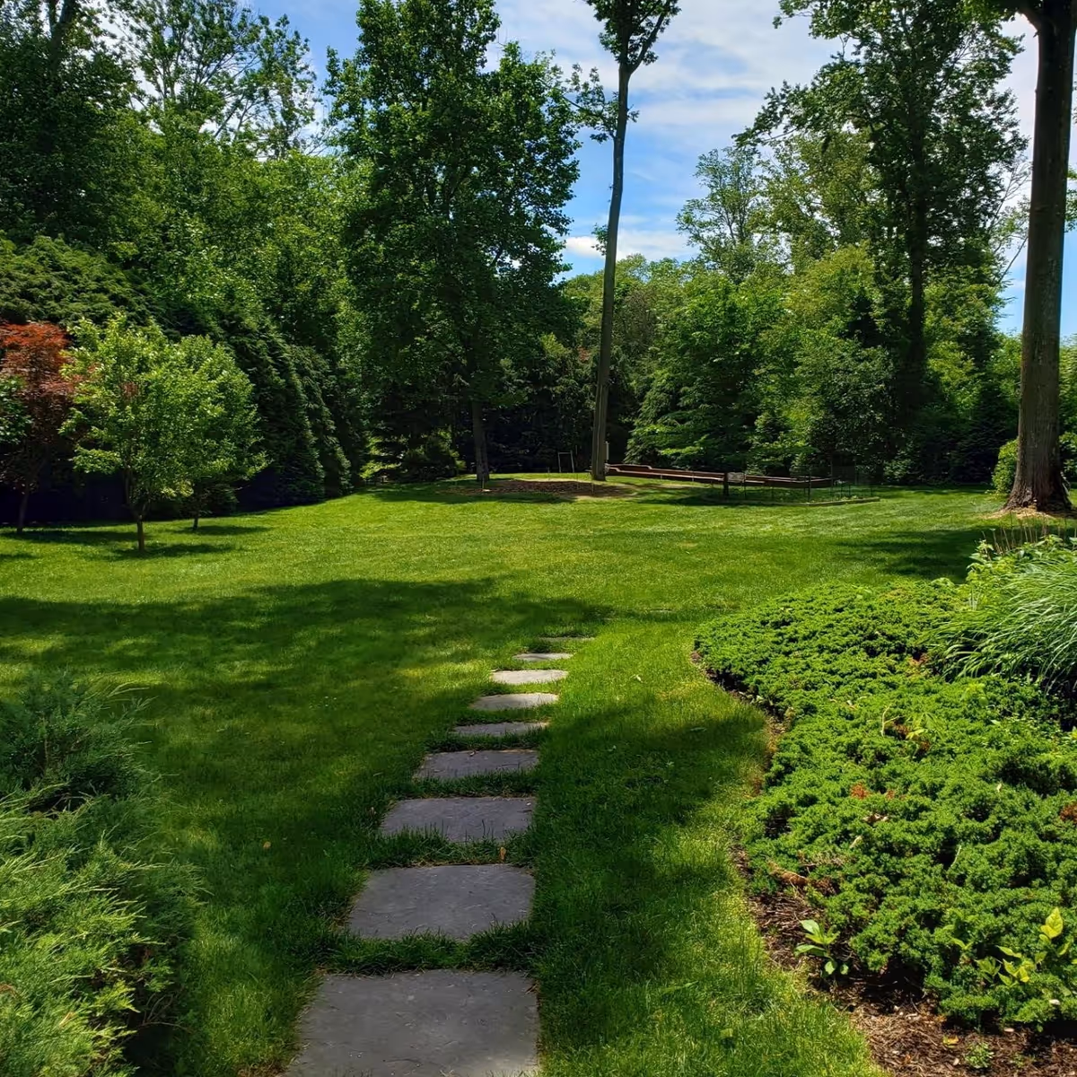 A lush green garden area with a stone pathway leading through well-maintained grass and surrounded by various trees and shrubs under a partly cloudy sky.