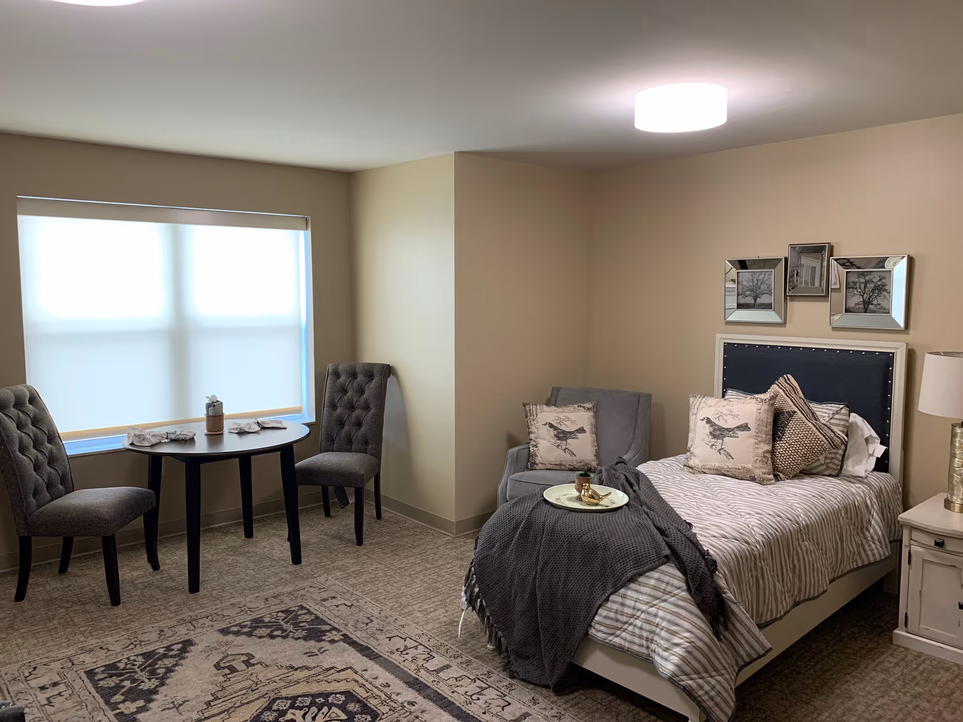 A furnished bedroom with a single bed, armchair, nightstand, small round table with two chairs, and a large window with blinds.