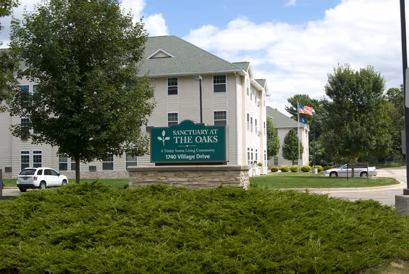 Exterior view of a senior living community building with beige siding and a green roof. In front of the building is a green sign that reads 'Sanctuary at The Oaks, A Trinity Senior Living Community, 1740 Village Drive'. There are trees, shrubs, a few parked cars, and an American flag on a flagpole near the building.