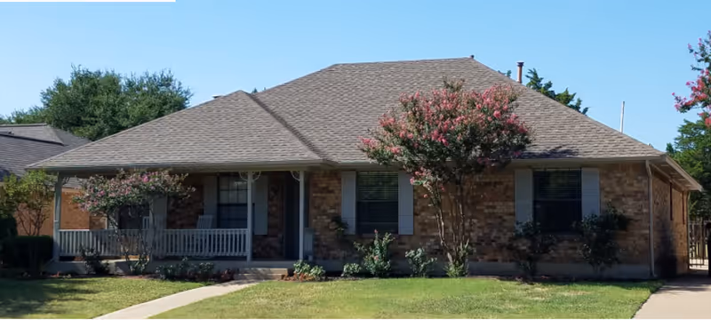 Single-story brick house with a covered front porch, flowering trees, and a neatly mowed lawn.