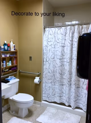 A bathroom with a toilet, a wall-mounted shelf holding toiletries, a grab bar with a toilet paper holder, a shower with a curtain featuring various cat illustrations, and a wall-mounted soap or paper towel dispenser. The wall has the text 'Decorate to your liking'.