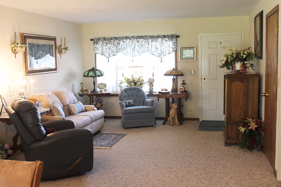 A cozy living room with a beige couch, a dark recliner chair, and a blue armchair in front of a window with floral curtains. There are two side tables with decorative lamps and floral arrangements, a wooden cabinet with flowers on top, and a small rug on the carpeted floor. The walls are light-colored with framed pictures and sconces.