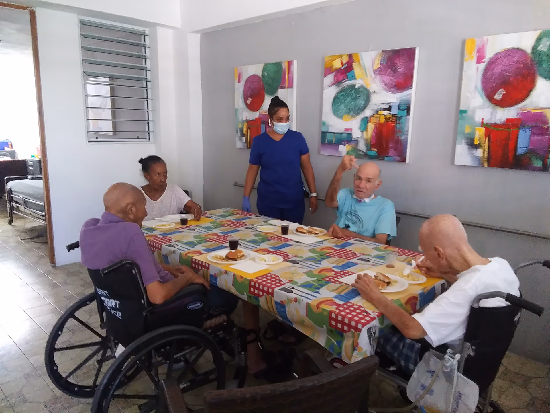 Several elderly residents in wheelchairs sit around a colorful table in a dining room while a caregiver stands nearby.