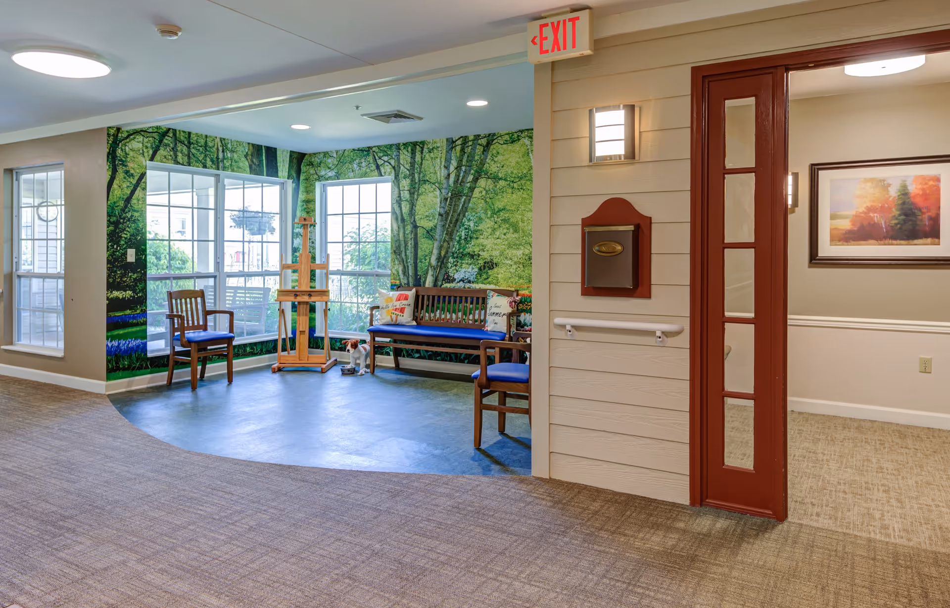Indoor seating area with wooden chairs and a bench with blue cushions, set against a wall mural depicting a lush green forest. There is an easel and a small dog figurine on the floor. The area is adjacent to a hallway with carpeted flooring, a red-framed glass door, a wall-mounted mailbox, and an exit sign above.