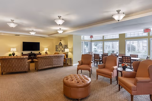 A spacious living room area in a senior living facility with multiple upholstered armchairs and sofas arranged around a large flat-screen TV mounted on the wall. The room has beige walls, carpeted floors, several table lamps, and large windows letting in natural light. There is a round leather ottoman in the center and a small table with a flower arrangement.