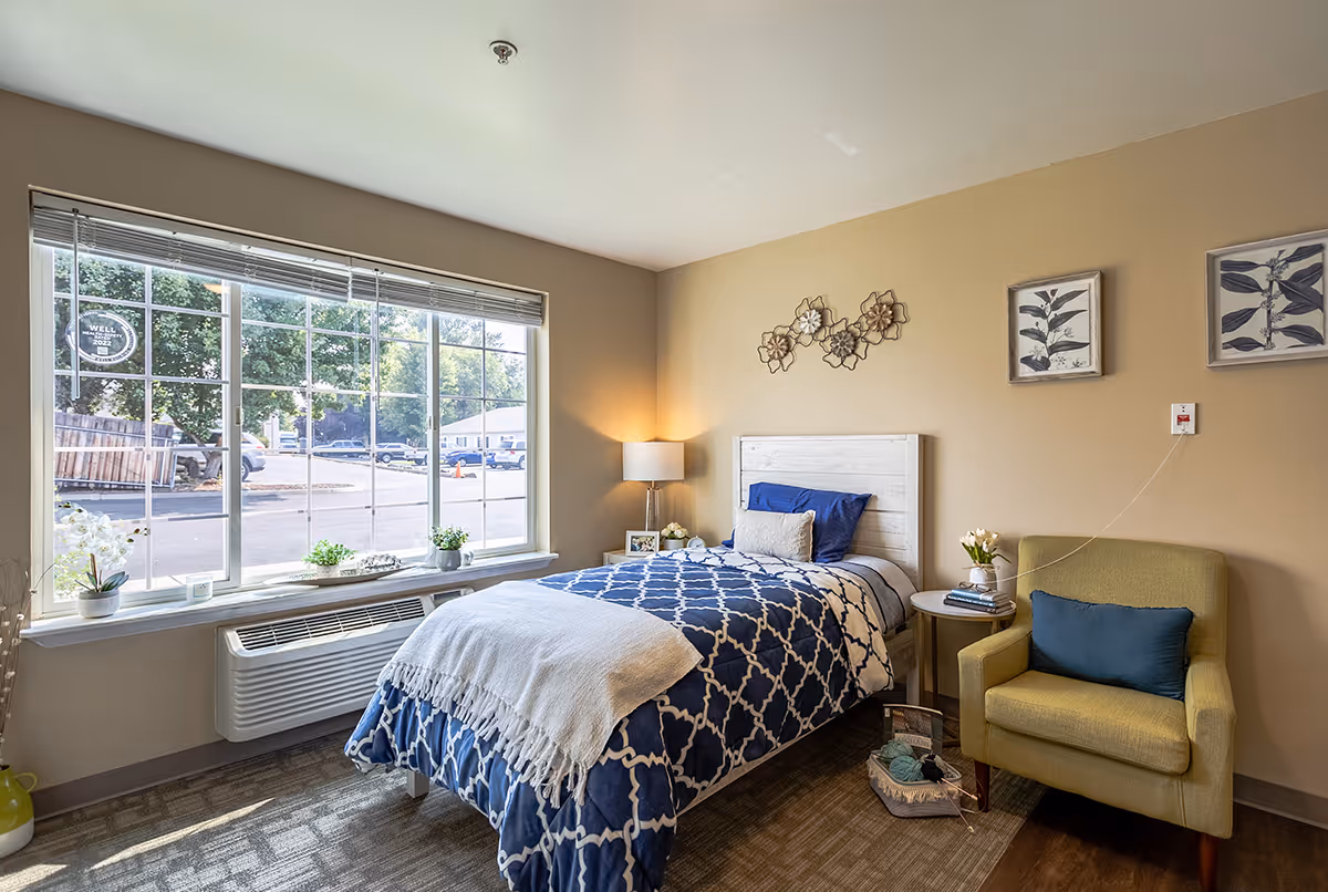 A cozy bedroom with a single bed covered in a blue and white patterned blanket and a white throw. Next to the bed is a small round table with books and a flower vase, and a green armchair with a blue cushion. The room has beige walls decorated with floral wall art and framed botanical prints. A large window with blinds lets in natural light and shows a parking lot and trees outside.
