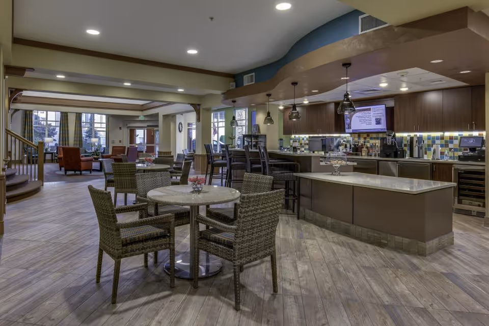 A spacious and modern common area in a senior living facility featuring a kitchen with a large island, bar seating with pendant lights, and multiple round tables with wicker chairs. In the background, there is a lounge area with armchairs and large windows allowing natural light to enter.