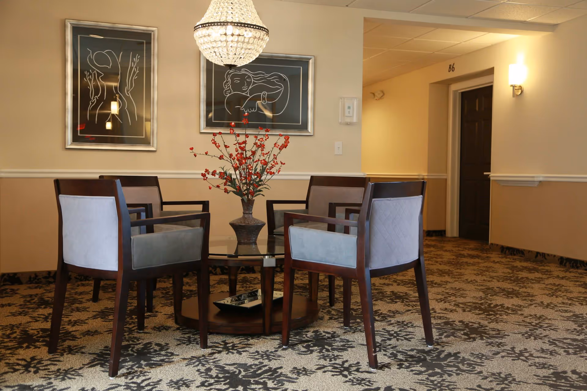 A seating area in a hallway with four wooden chairs with gray cushions arranged around a round glass-top table holding a vase with red flowers. Two framed abstract line art pictures hang on the beige wall behind the chairs, and a crystal chandelier hangs from the ceiling. A dark door labeled 'B6' is visible in the hallway.