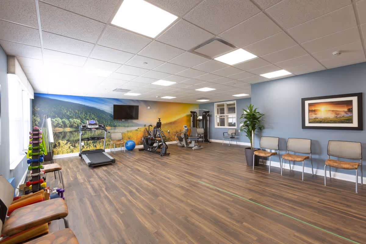 A spacious fitness room with wooden flooring and light blue walls. The room contains exercise equipment including a treadmill, stationary bike, and weight rack with colorful dumbbells. Several chairs are lined up against the walls, and a large mural of a scenic landscape with trees and a lake covers one wall. A flat-screen TV is mounted on the mural wall, and a framed picture of a sunset hangs on another wall. The room is well-lit with ceiling lights and natural light from windows.