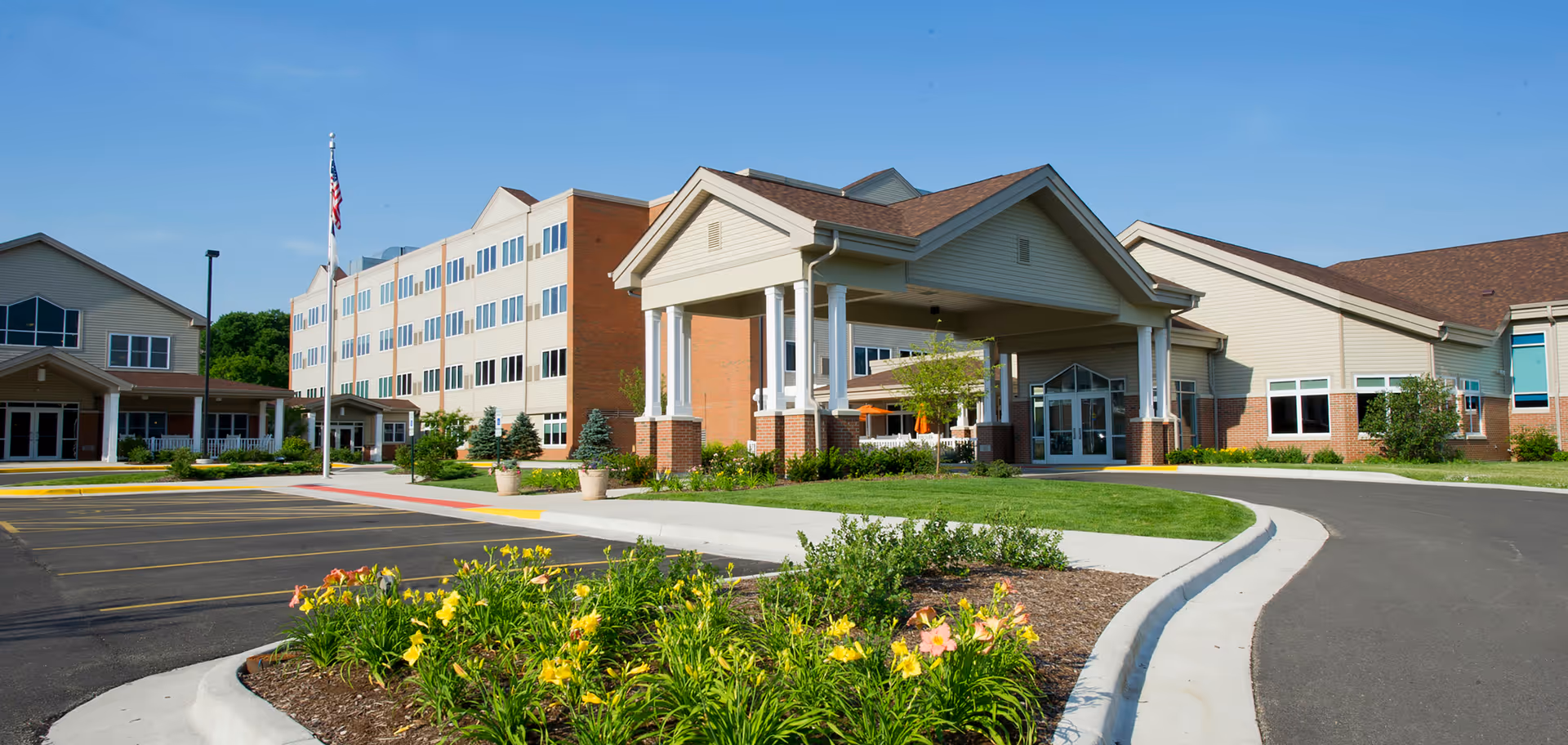 Main entrance of Pleasant View Lutheran Home with a porte-cochere, landscaped flowerbeds, and a multi-story brick-and-siding building under a blue sky.