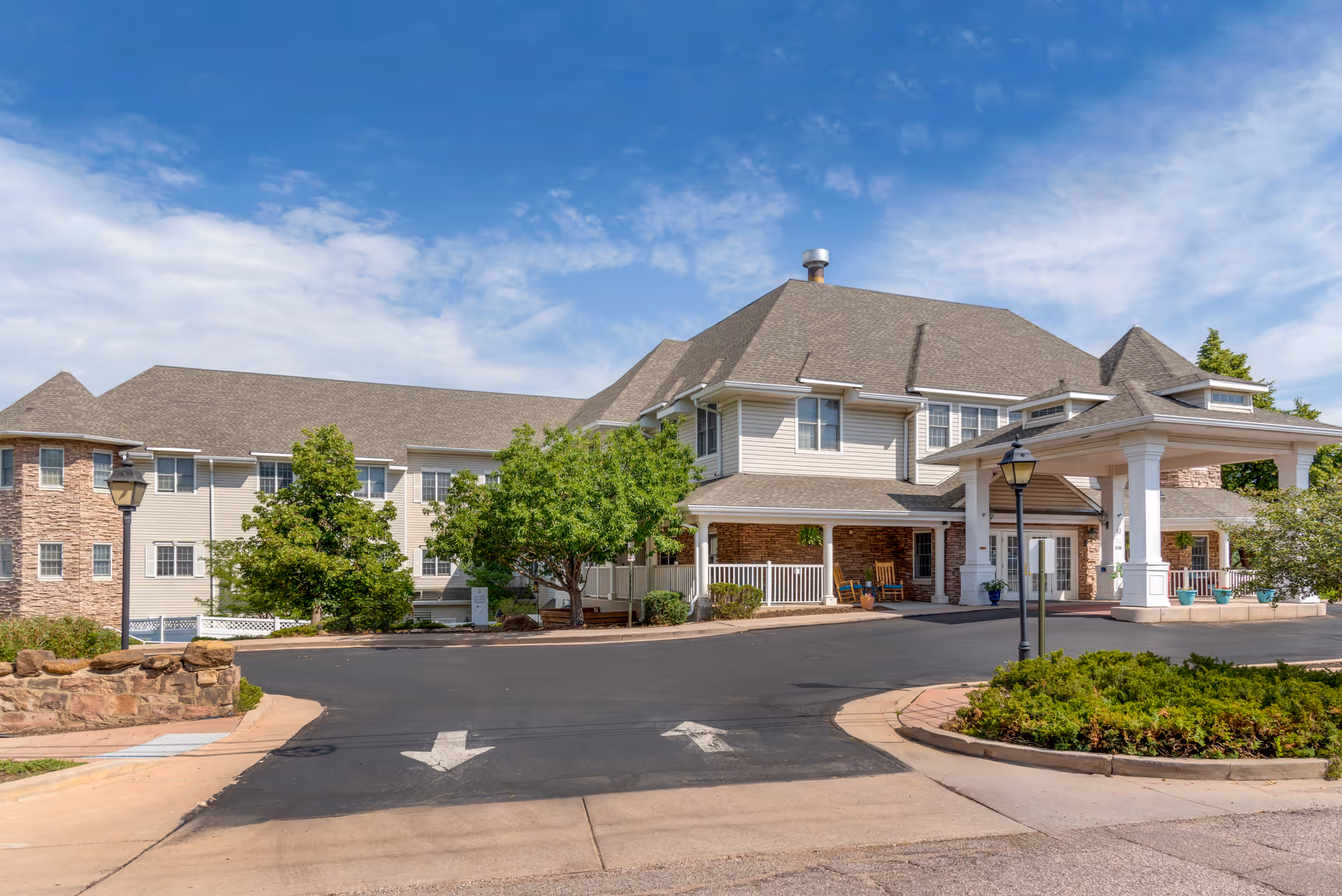 Front exterior of a senior living building with a covered porte-cochere entrance, circular driveway with arrows, and landscaping under a blue sky.