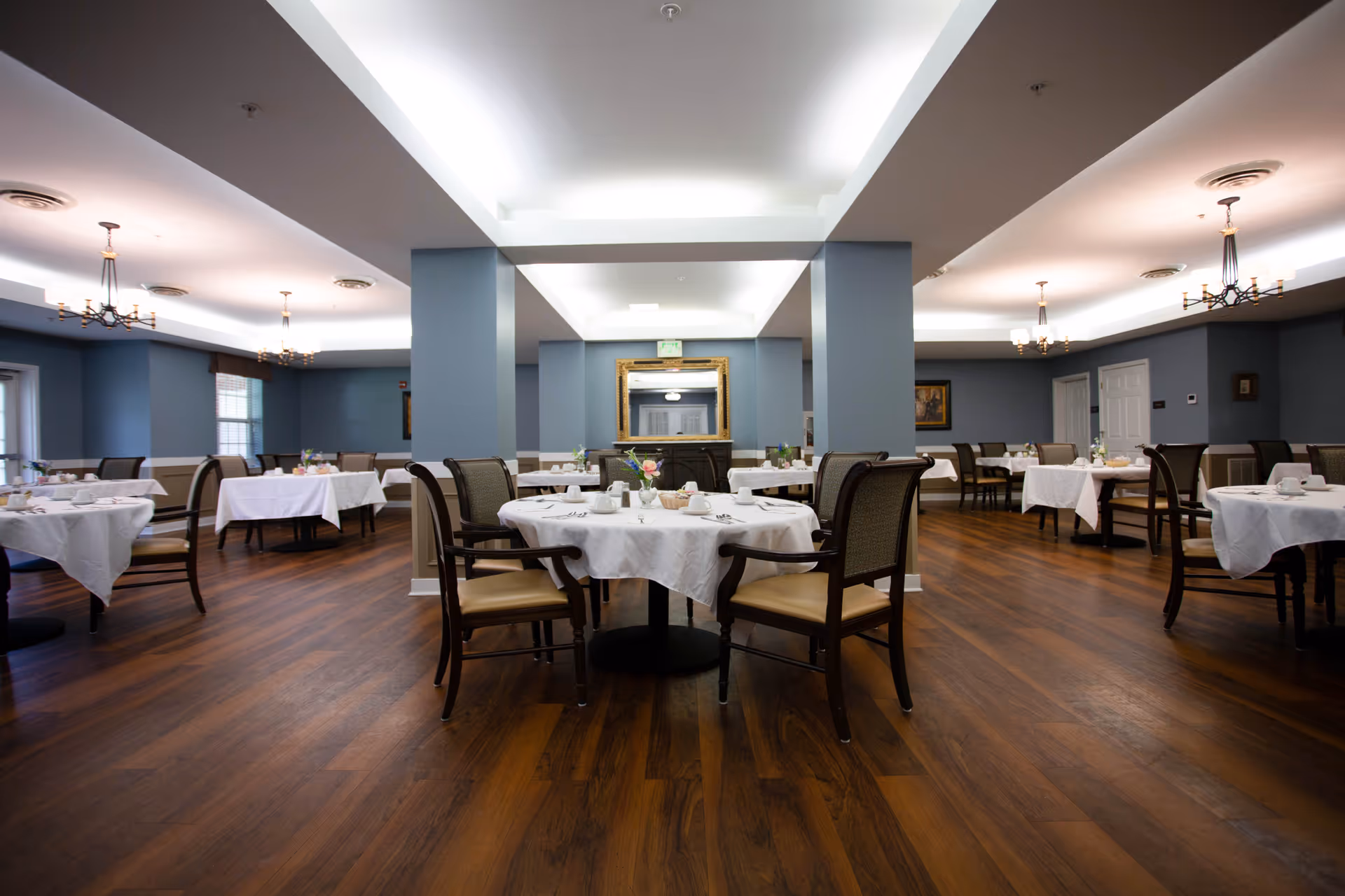 A spacious dining room with multiple round tables covered with white tablecloths, each set with cups, plates, and small flower arrangements. The room has wooden flooring, blue-gray walls, and several chandeliers hanging from the ceiling. There is a large mirror on the far wall between two columns.