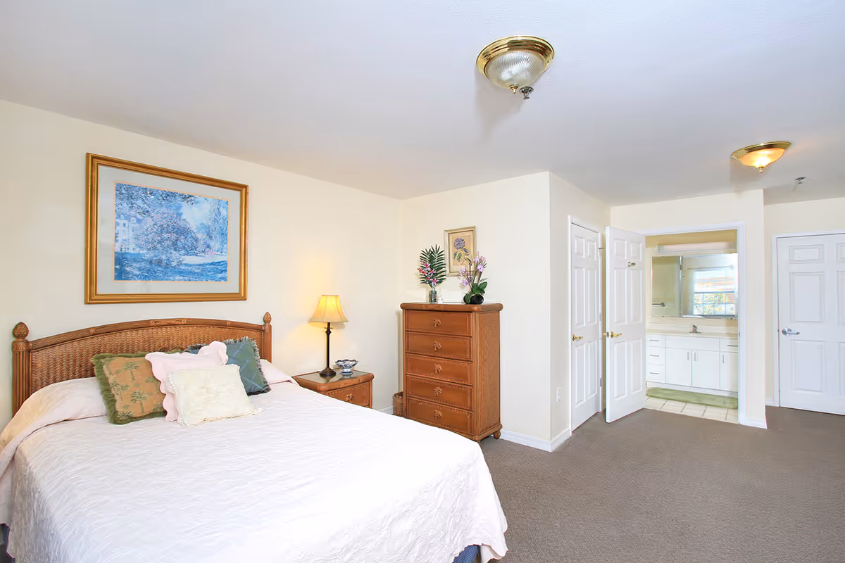 A bright bedroom with a large bed covered in a white quilt and several decorative pillows. There is a wooden nightstand with a lamp and a wooden dresser with flowers on top. A framed painting hangs on the wall above the bed. The room has beige walls and carpeted floor. An open door reveals a bathroom with a large mirror and white cabinetry.
