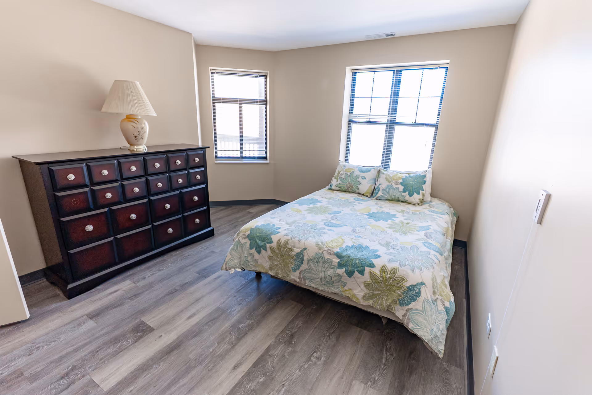 A small bedroom with a double bed covered in a floral patterned bedspread in shades of blue and green. There are two matching pillows on the bed. The room has two windows with blinds, light beige walls, and wood-look flooring. A dark wooden dresser with multiple drawers and a decorative lamp on top is positioned against one wall.
