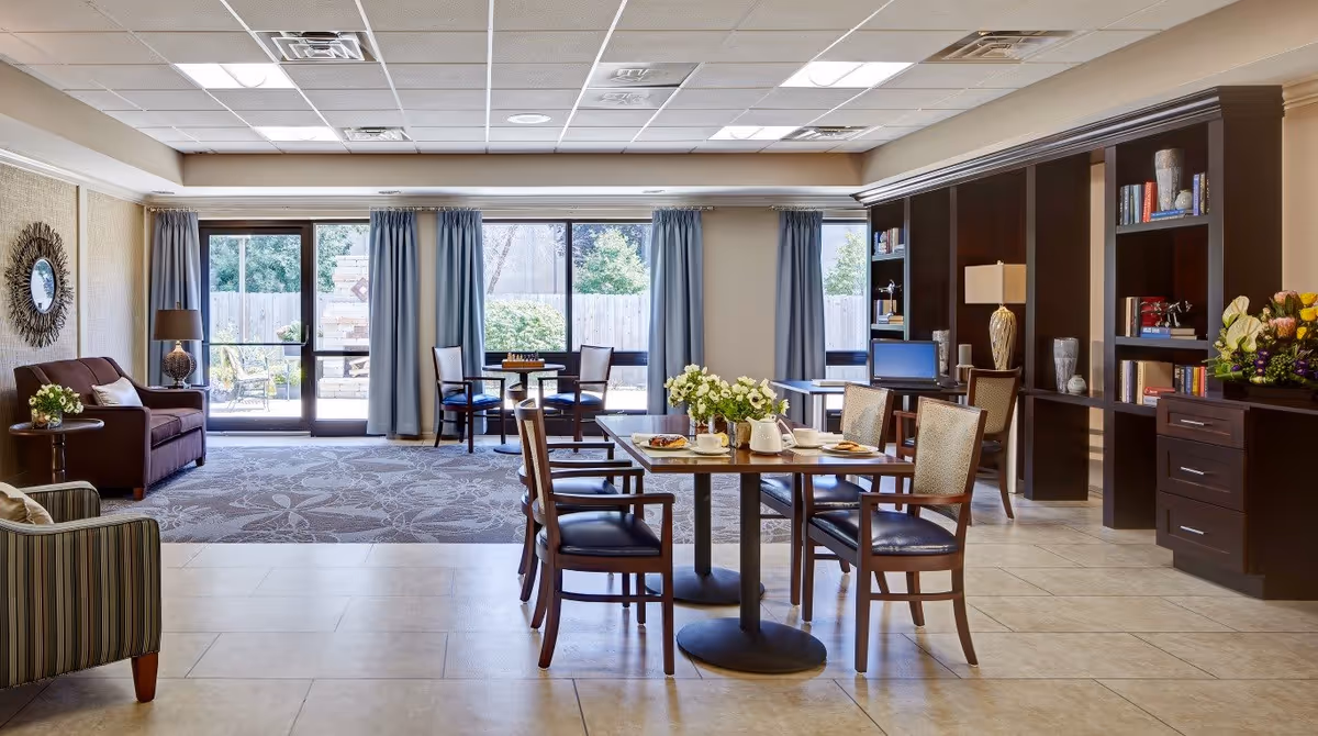 Bright communal dining and lounge room with tables and chairs, sofas, bookshelves, and large windows overlooking an outdoor area.