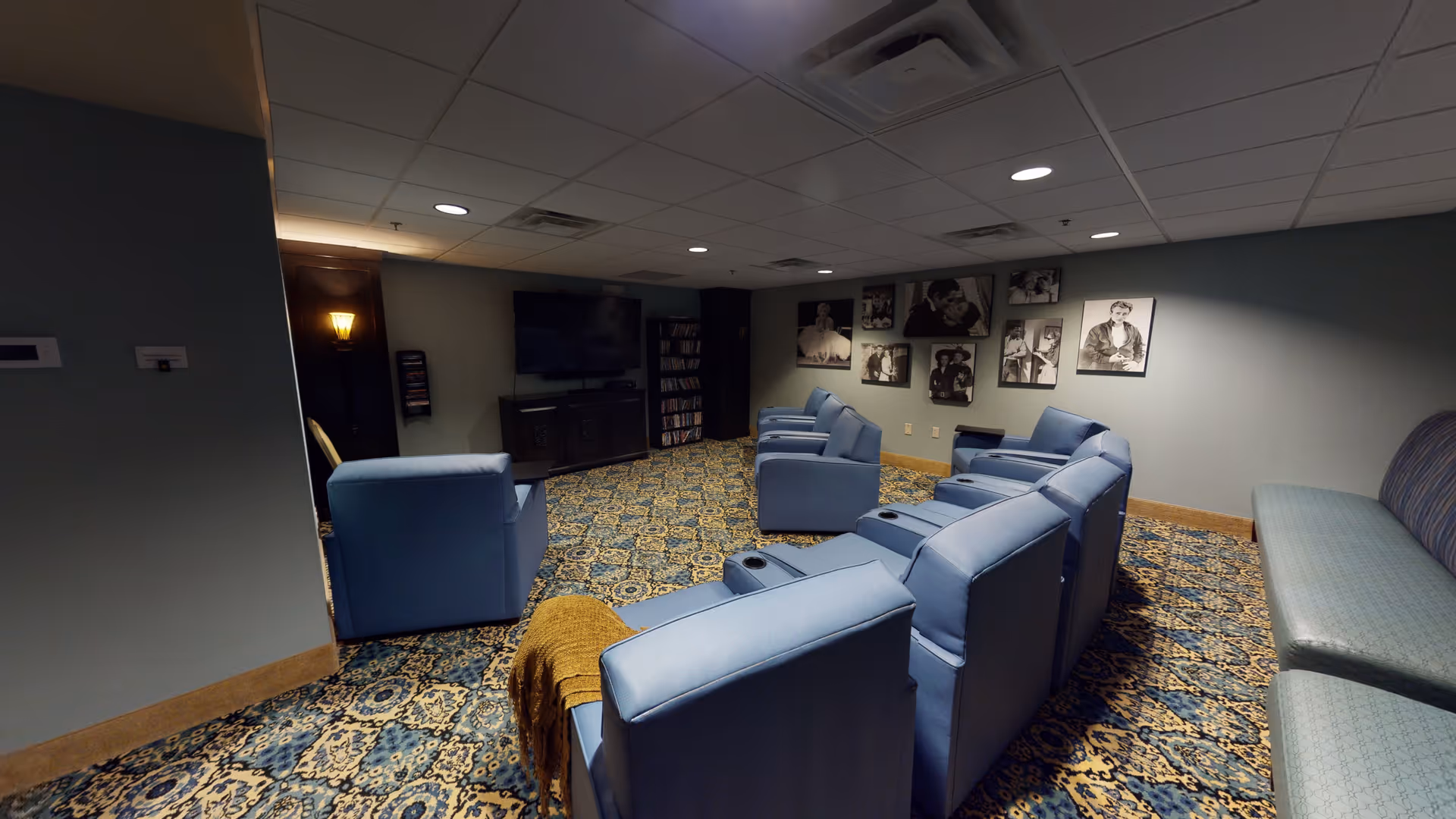 A cozy common room with multiple blue recliner chairs arranged in rows facing a large flat-screen TV mounted on a dark wooden cabinet. The room has patterned carpet flooring and soft lighting. Black and white framed photos are displayed on the wall behind the chairs. There is also a long cushioned bench along one wall.