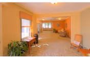 A long carpeted communal living area with upholstered chairs, side tables, plants, and windows.
