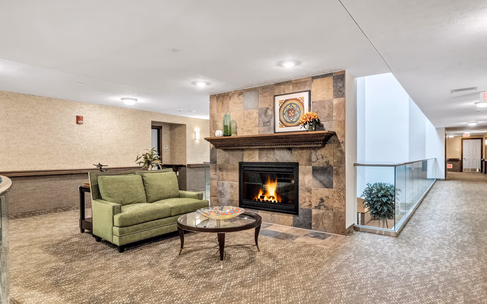 A cozy indoor seating area in a retirement community featuring a green upholstered loveseat, a round glass coffee table with a decorative bowl, and a lit fireplace with a tiled surround and a wooden mantel. Above the mantel is a framed piece of artwork and decorative items including a vase and flowers. The space has carpeted floors, beige walls, recessed lighting, and a hallway with glass railing leading to other rooms.