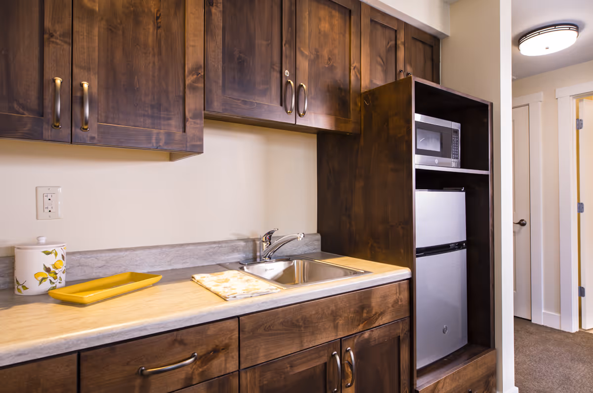A small kitchen area with dark wooden cabinets, a countertop with a sink, a yellow tray, and a decorative container. There is a microwave and a mini refrigerator built into a wooden cabinet. The area is well-lit with a ceiling light and a hallway with doors is visible in the background.