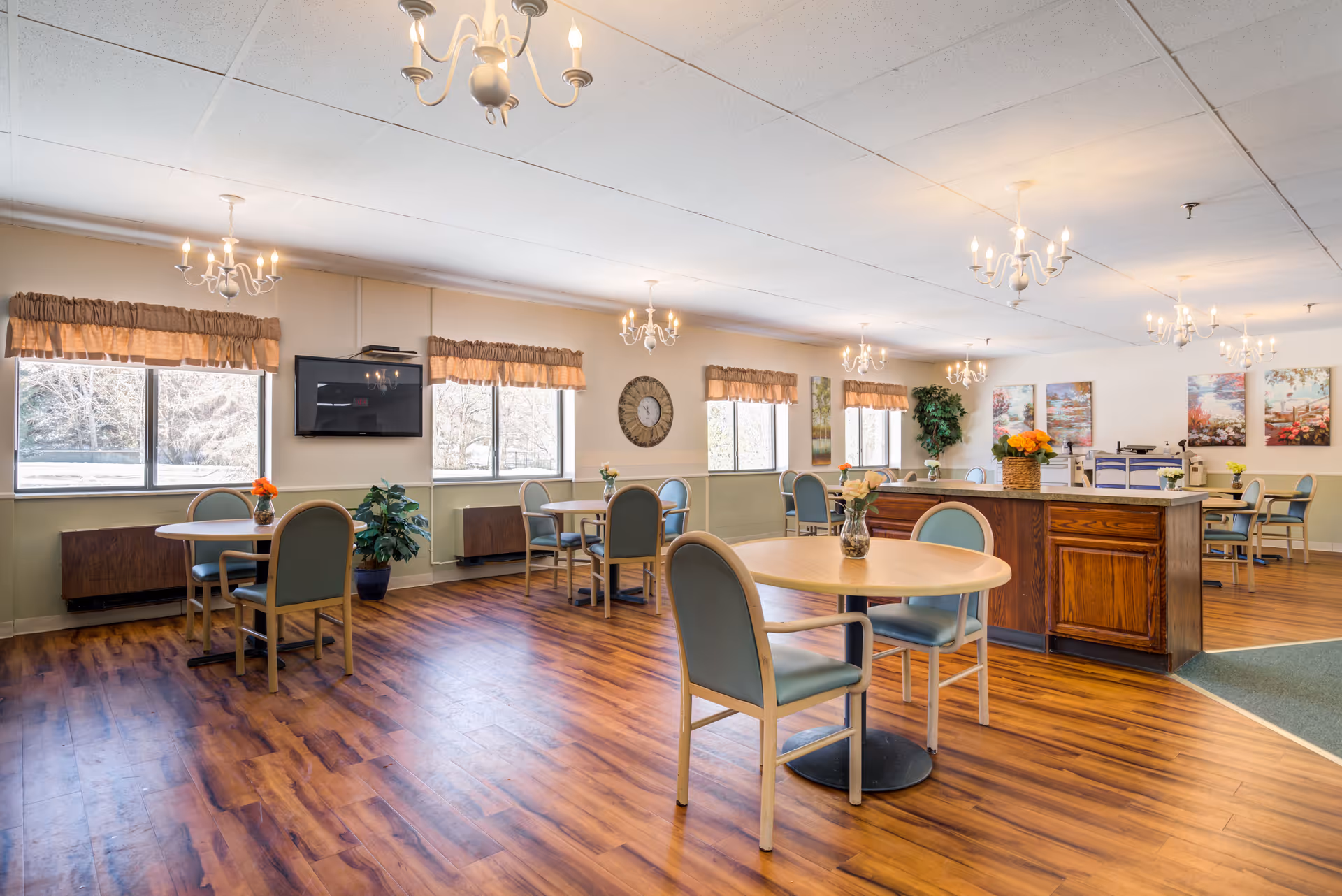 A spacious dining room in an assisted living facility with several round tables and chairs arranged neatly. The room has large windows with valance curtains allowing natural light to fill the space. There are chandeliers hanging from the ceiling, a wall-mounted TV, a large clock, and decorative paintings on the walls. The floor is wood laminate, and there are small flower arrangements on each table.