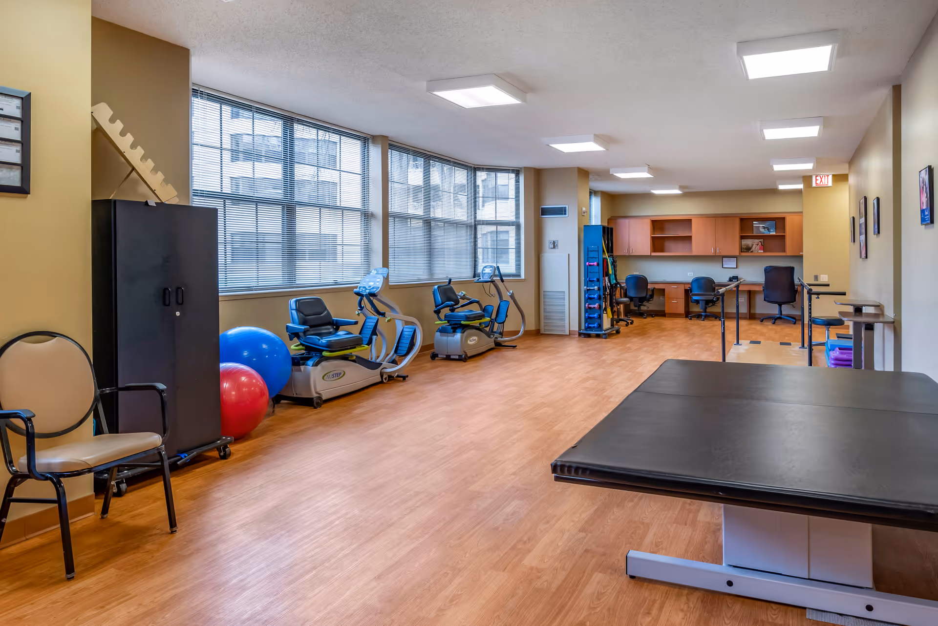 A spacious rehabilitation or physical therapy room with exercise equipment including stationary recumbent bikes, therapy balls, and parallel bars. There are chairs along the left wall, large windows letting in natural light, wooden flooring, and a black padded therapy table in the foreground. The back wall has wooden cabinets and a desk area with chairs.