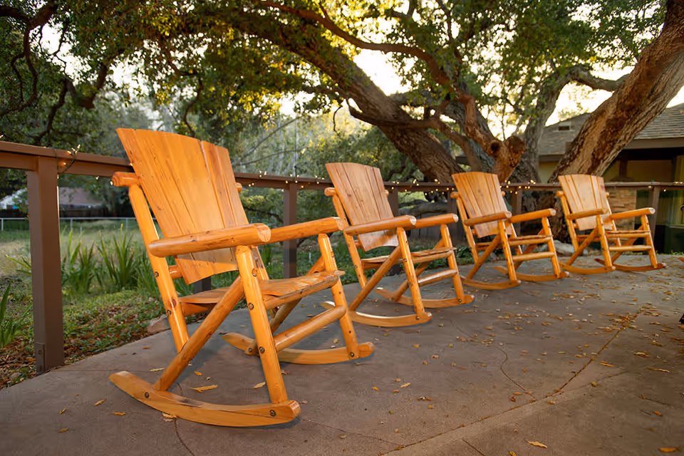 Four wooden rocking chairs arranged in a row on a concrete patio with a wooden railing and large trees in the background during daylight.