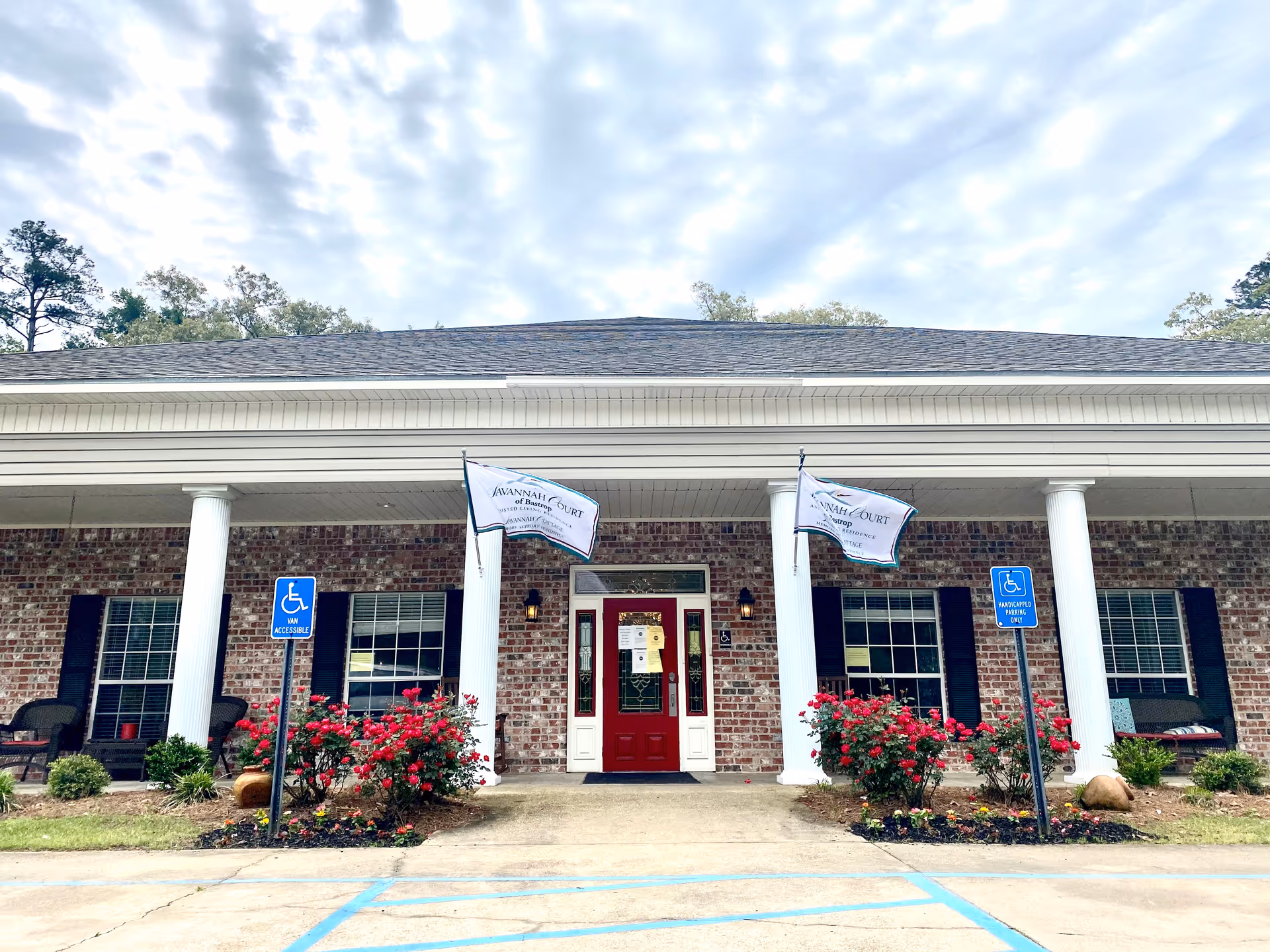 Front exterior view of a brick building with white columns and a red door. There are two flags with the text 'Savannah Court' hanging between the columns. Two handicap parking signs are visible in front of the building, along with flower beds containing red flowers and greenery. The sky is partly cloudy.