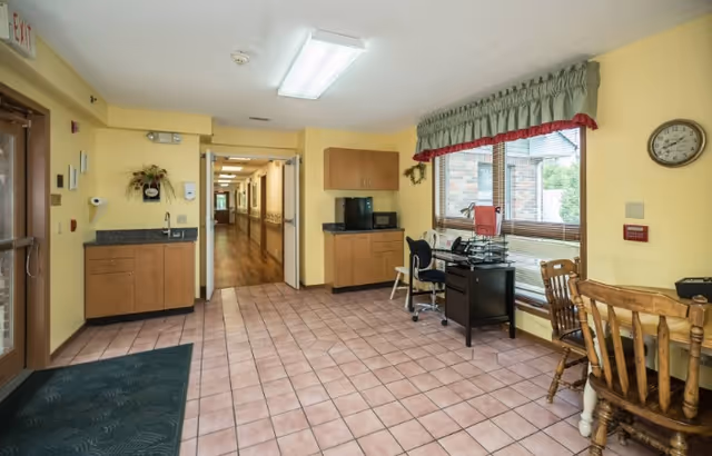 Interior view of a senior living facility reception or common area with tiled floor, yellow walls, wooden cabinets, a desk with office supplies, chairs, a large window with green and red valance, and a hallway leading further inside the building.