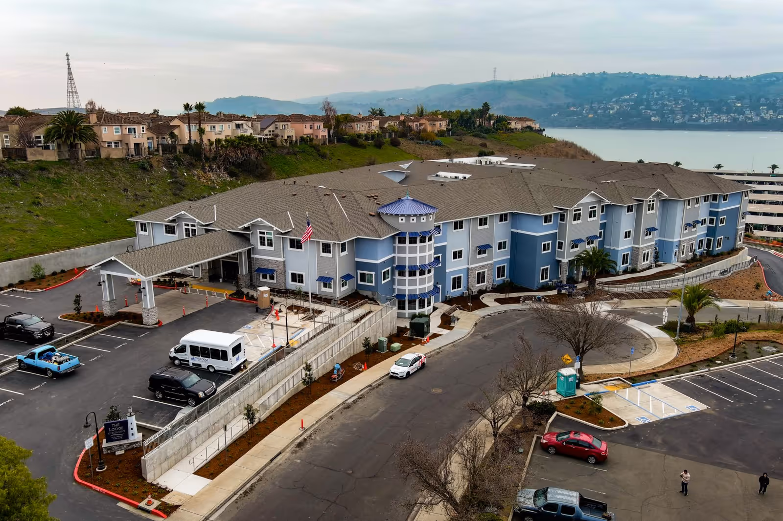 Aerial view of The Lodge at Glen Cove, a large multi-story senior living facility with a blue and white exterior. The building is situated near a body of water with hills and houses in the background. The parking lot in front has several vehicles parked, and there is a covered entrance with an American flag. The surrounding area includes landscaped greenery and a few trees.
