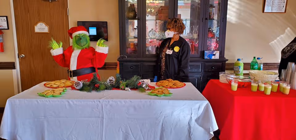 A festive indoor scene with a person dressed as the Grinch in a Santa outfit standing behind a table covered with a white tablecloth. The table has plates of cookies and holiday decorations. Another person wearing a face mask and gloves stands behind the table, next to a black cabinet with glass doors. To the right, there is a red tablecloth-covered table with drinks and cups.