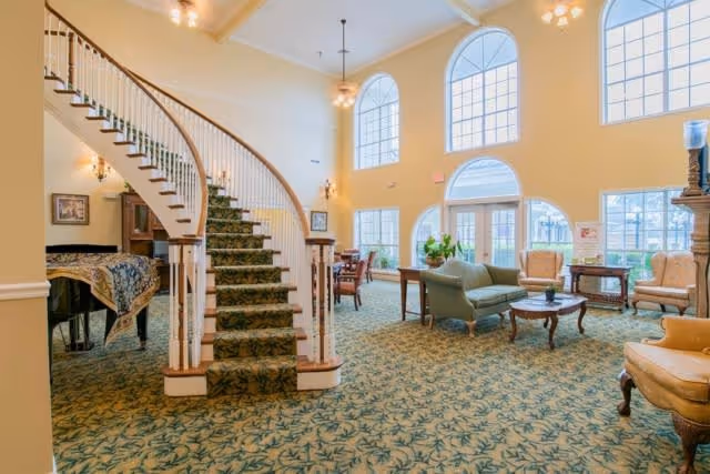 A spacious and well-lit living room area with large arched windows allowing natural light to fill the space. The room features a curved staircase with a patterned carpet, several upholstered chairs, a sofa, wooden tables, and a piano covered with a decorative cloth. The carpet has a green leafy pattern, and the walls are painted a soft yellow.