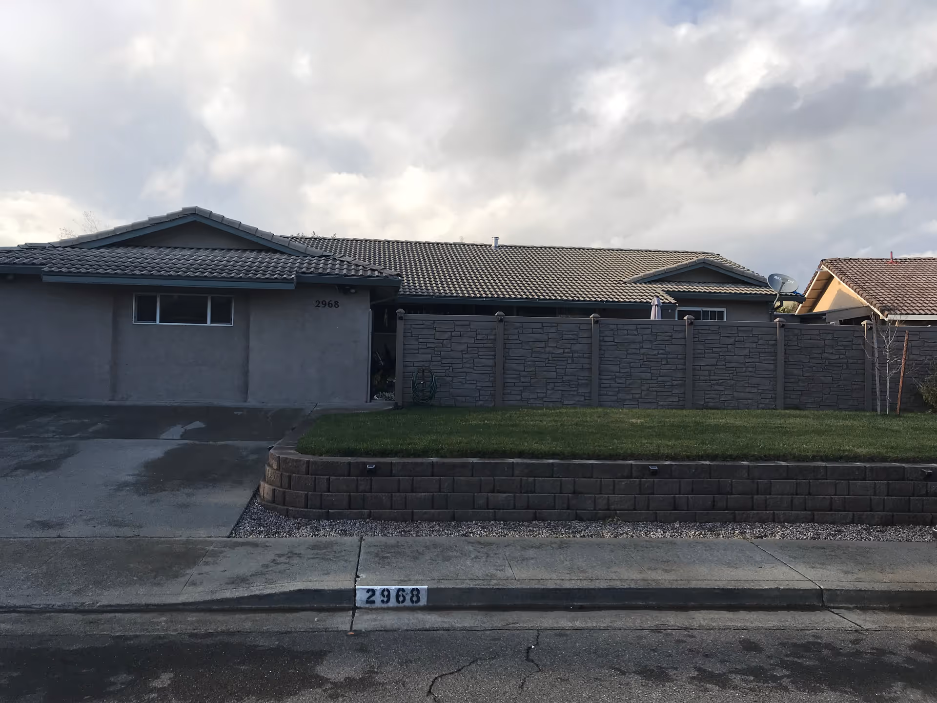 Exterior front view of a single-story residential building with a tiled roof, a driveway, a small lawn bordered by a stone retaining wall, and a tall privacy fence. The house number 2968 is visible on the curb and the wall.