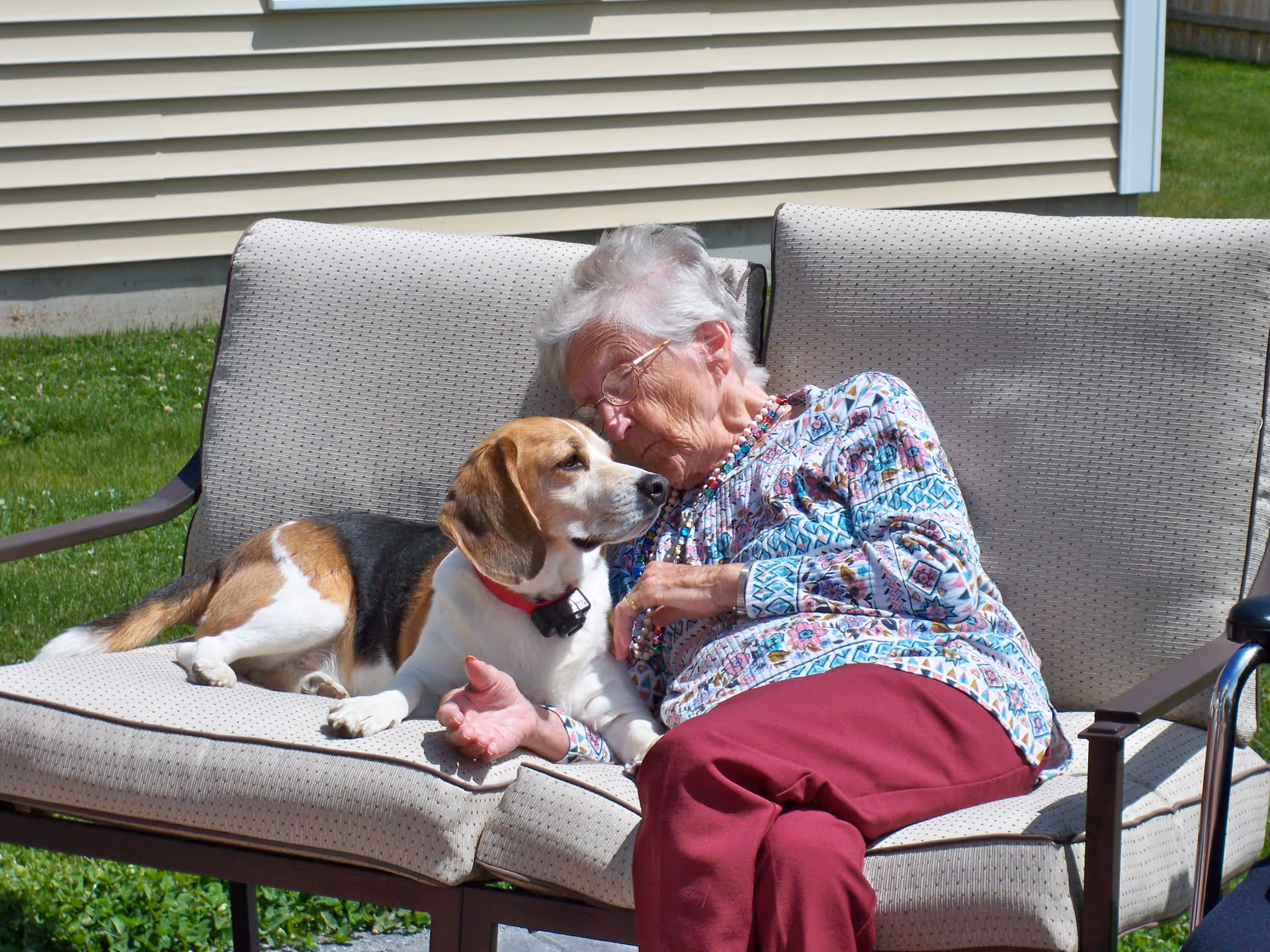 An elderly woman with white hair and glasses is sitting on an outdoor cushioned bench, affectionately leaning her head against a beagle dog lying next to her. The background shows the side of a beige house and green grass.