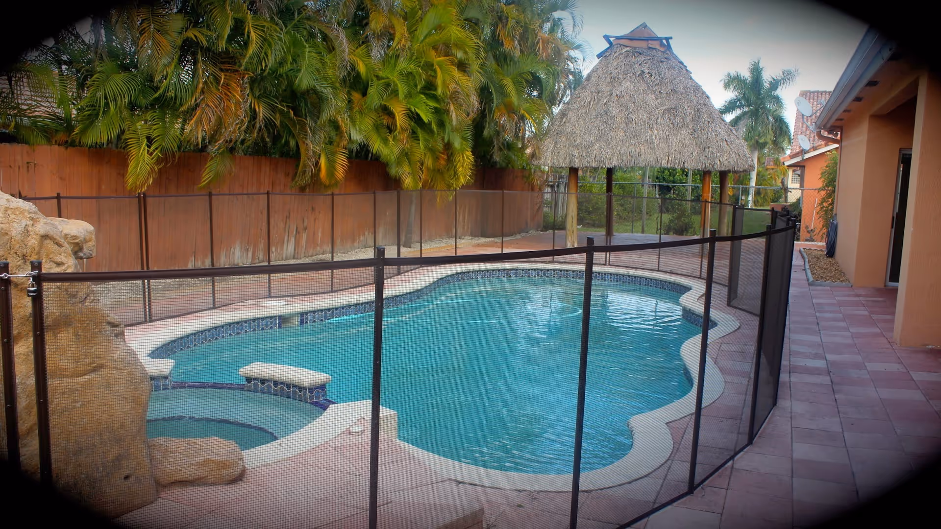 Outdoor swimming pool area with a safety fence around the pool. There is a small attached hot tub, a rock feature, palm trees, and a thatched roof gazebo in the background. The pool is surrounded by a tiled patio and a wooden fence with greenery behind it.