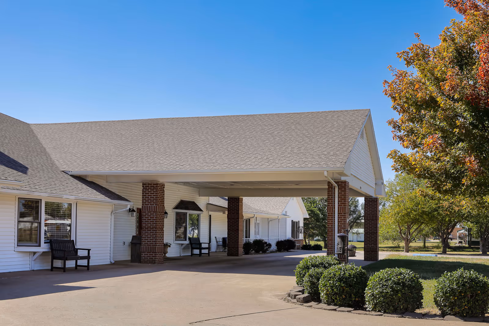 Exterior view of Homestead Assisted Living of Russell showing a covered entrance with brick columns, white siding, benches near windows, and well-maintained landscaping including trimmed bushes and trees with autumn foliage under a clear blue sky.