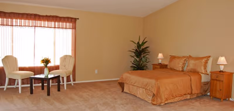 A spacious bedroom with beige walls and carpet. The room features a bed with orange bedding and two matching pillows, flanked by two wooden nightstands each with a small lamp. There is a green potted plant next to the bed. Near a large window with sheer curtains, there are two cream-colored chairs and a small dark wooden table with a vase of flowers.