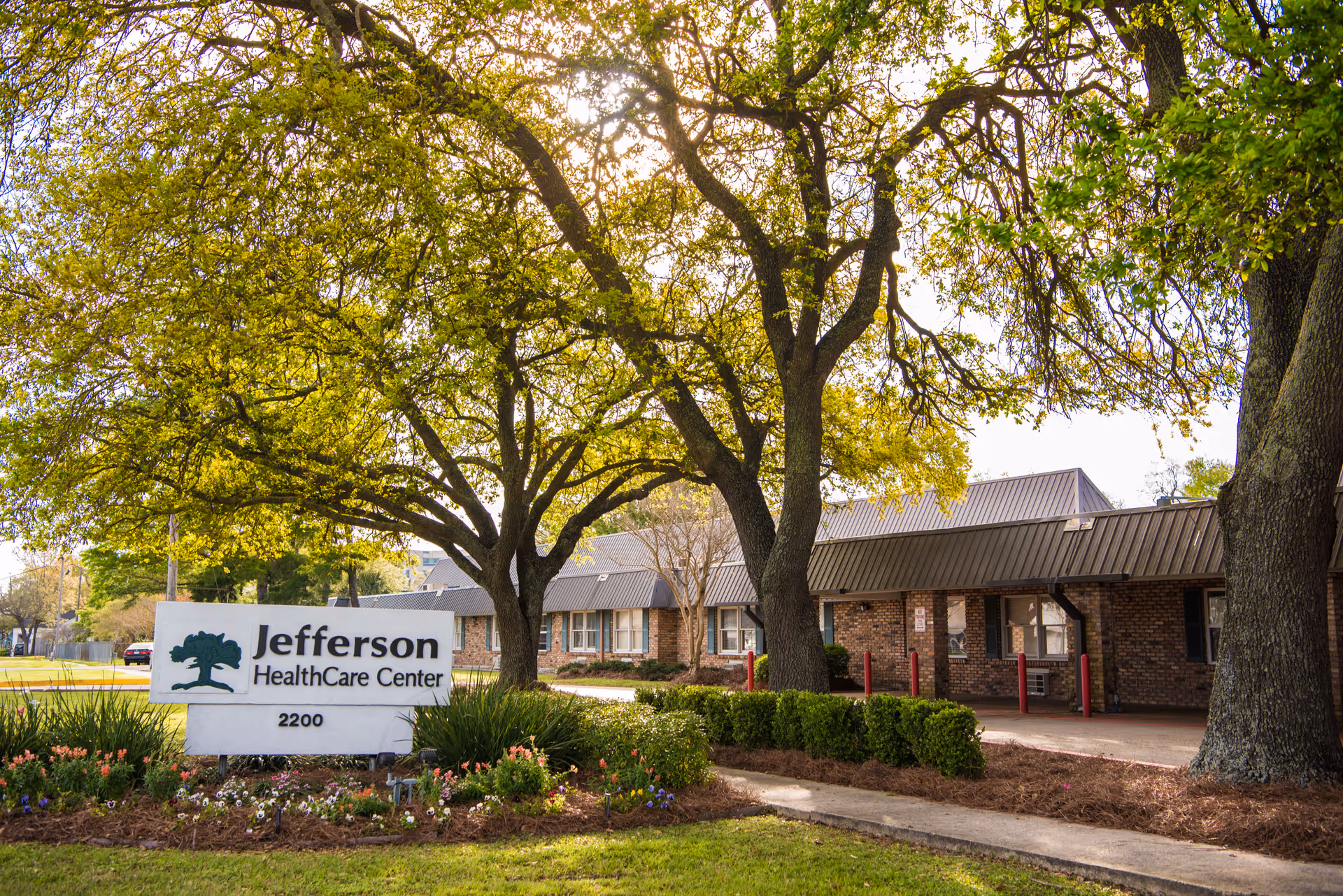 Sign for Jefferson HealthCare Center in front of a single-story brick building with large trees and landscaped flowerbeds.