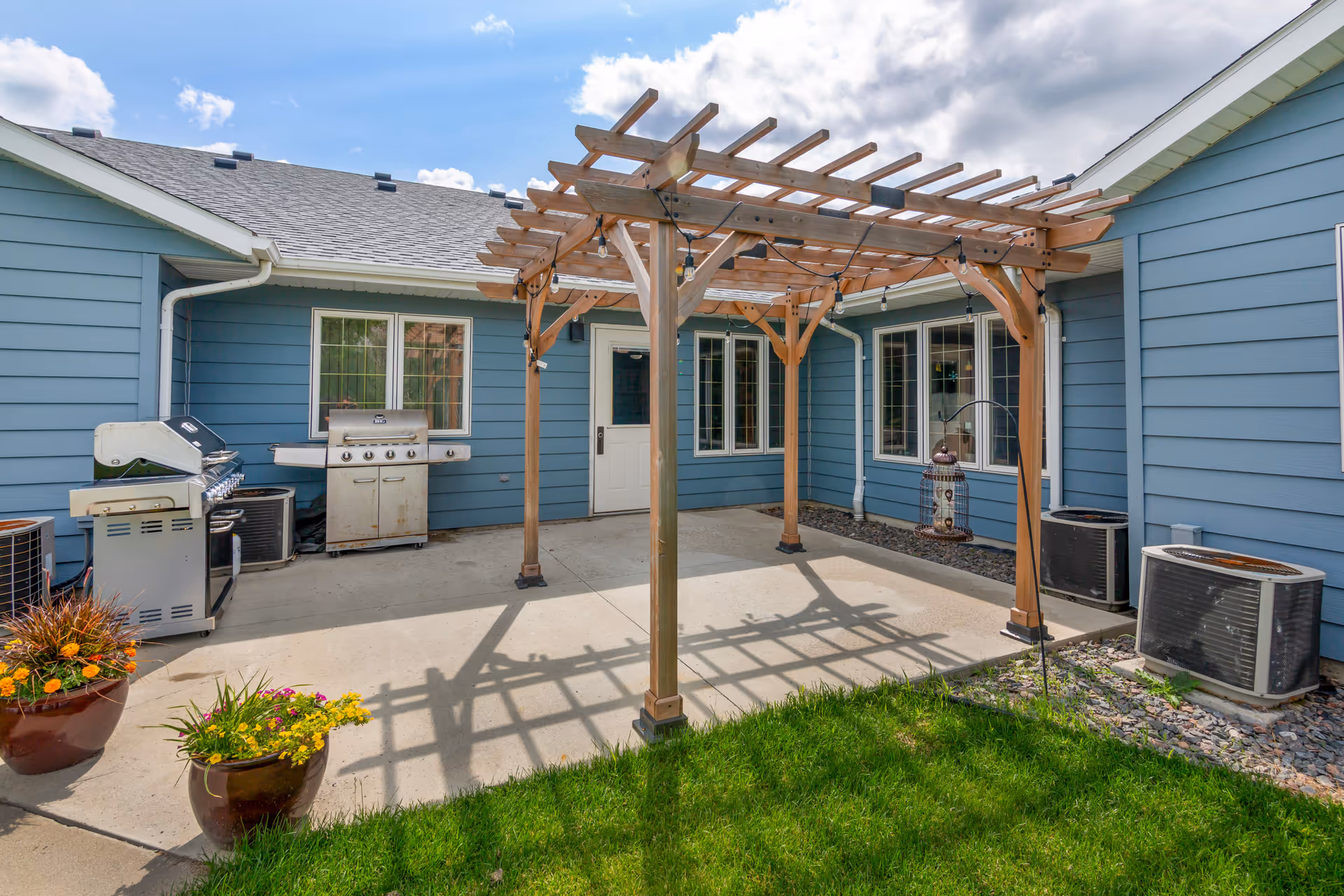 Patio courtyard with a wooden pergola, barbecue grills, potted flowers, and blue-sided building walls.