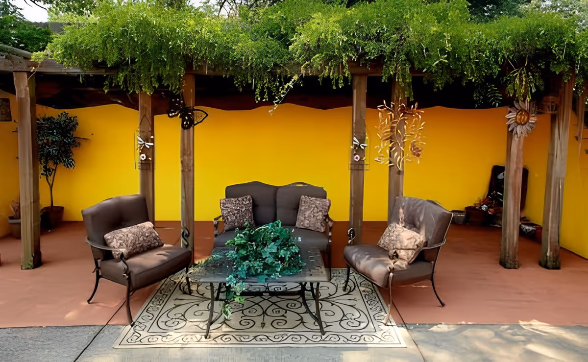 Outdoor covered patio with a loveseat and three cushioned chairs arranged around a coffee table under a pergola in front of a bright yellow wall.
