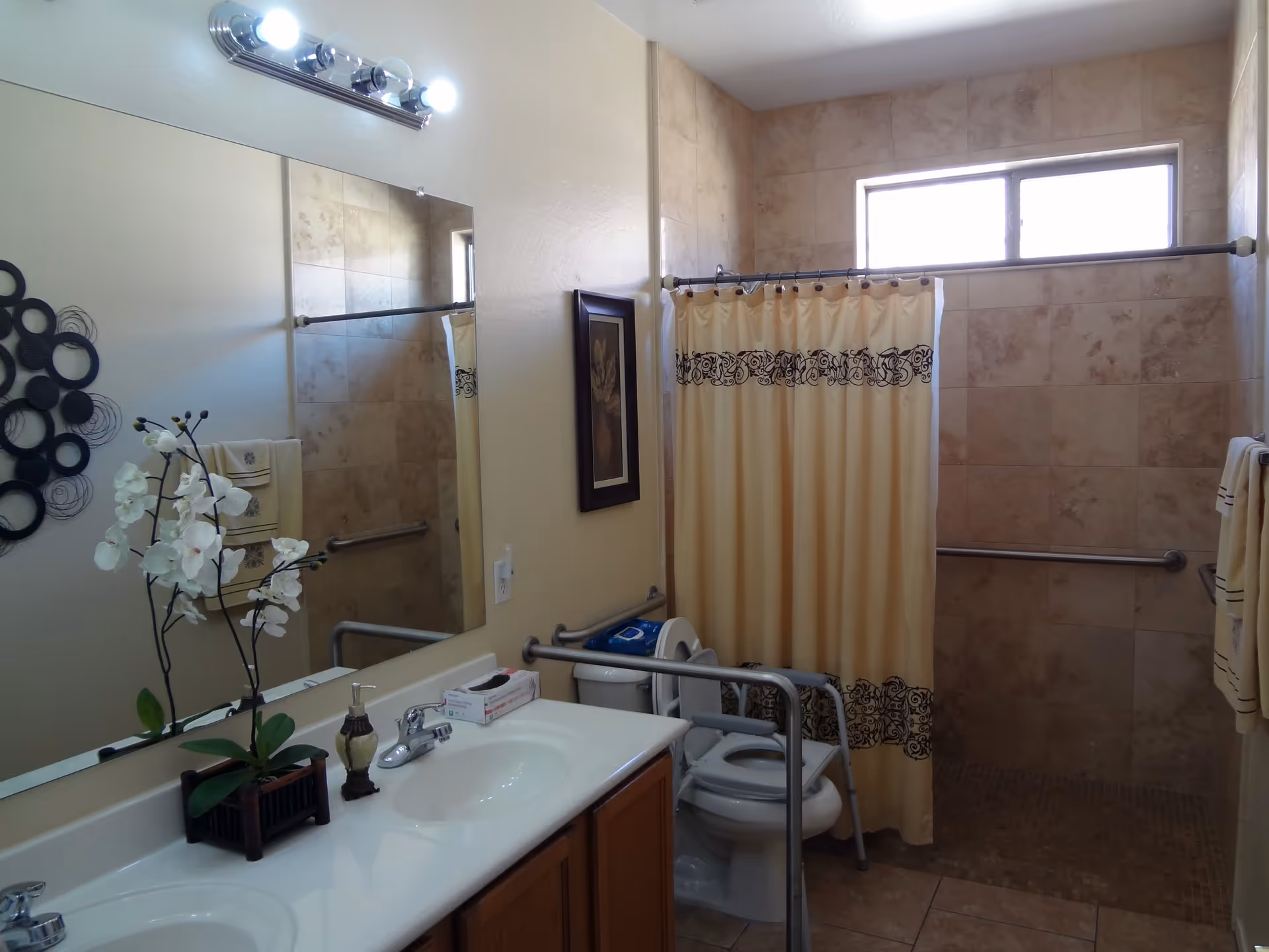 Accessible bathroom with a double-sink vanity and mirror, toilet with safety rails, and a tiled walk-in shower with a curtain and grab bars.