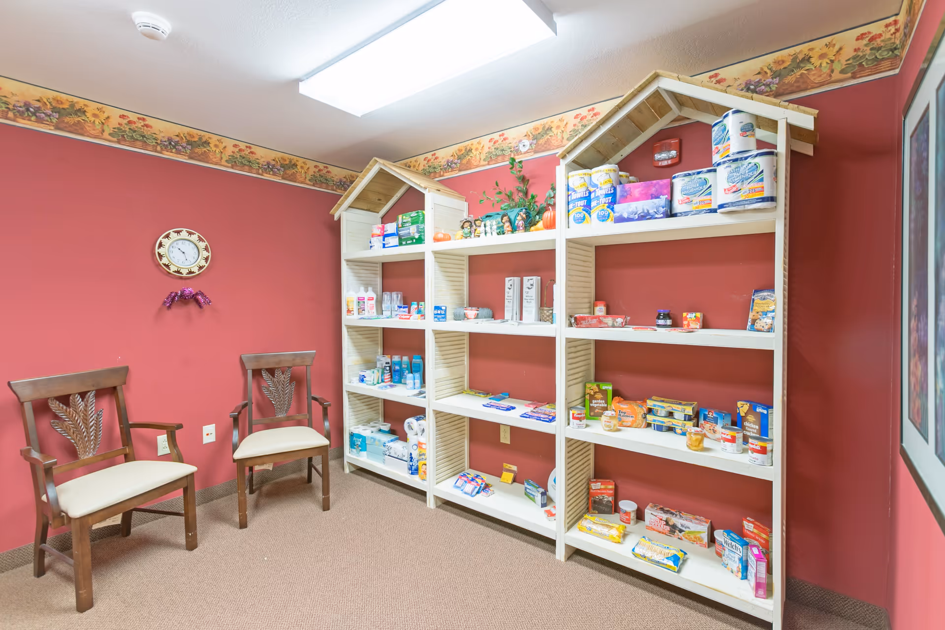 A room with red walls and beige carpet featuring two wooden chairs with cream cushions on the left. On the right, there are white shelves shaped like small houses filled with various packaged food items and household supplies. A round wall clock is mounted on the wall above the chairs, and a floral border runs along the top of the walls.