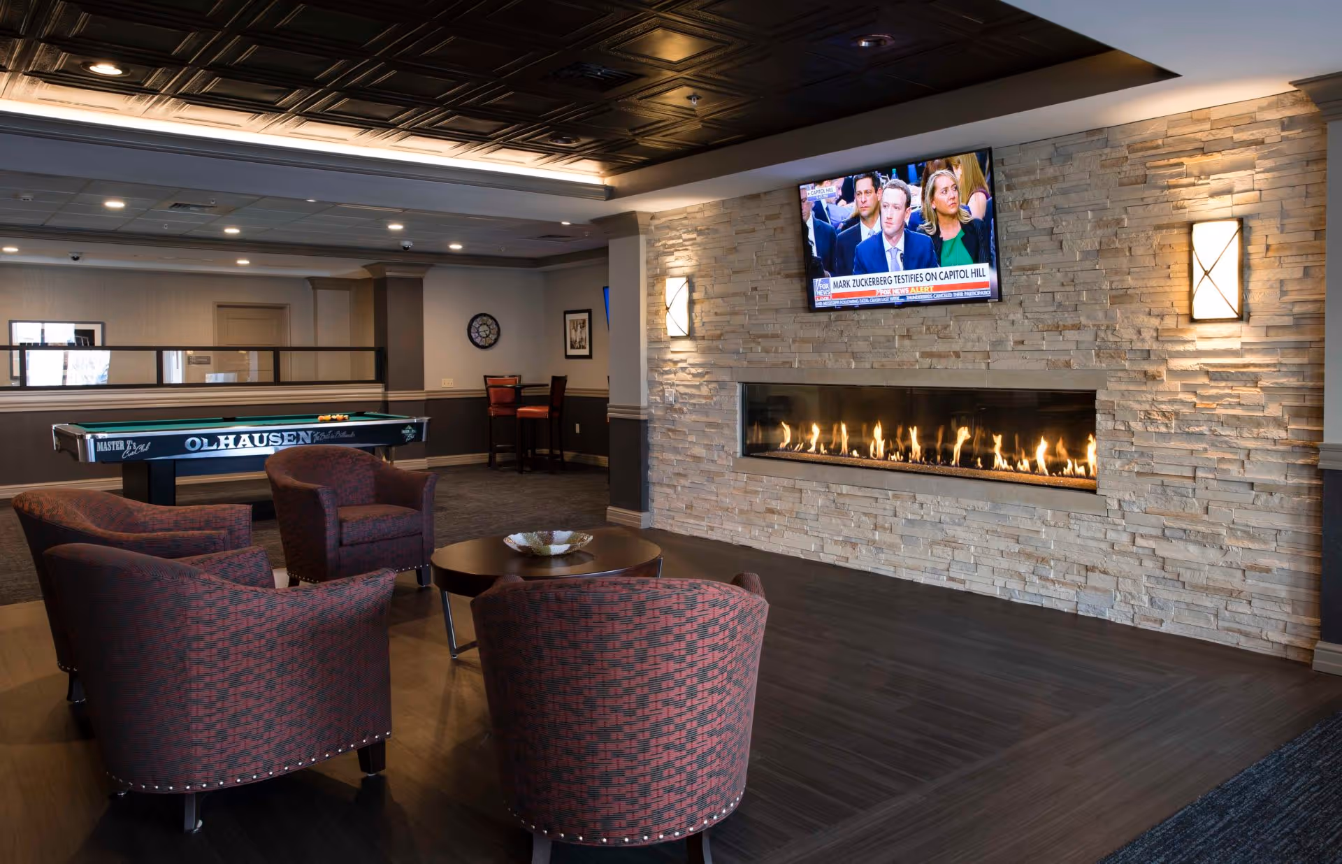 A cozy communal lounge with upholstered chairs around a coffee table, a long gas fireplace set in a stone wall, a wall-mounted TV, and a pool table in the background.