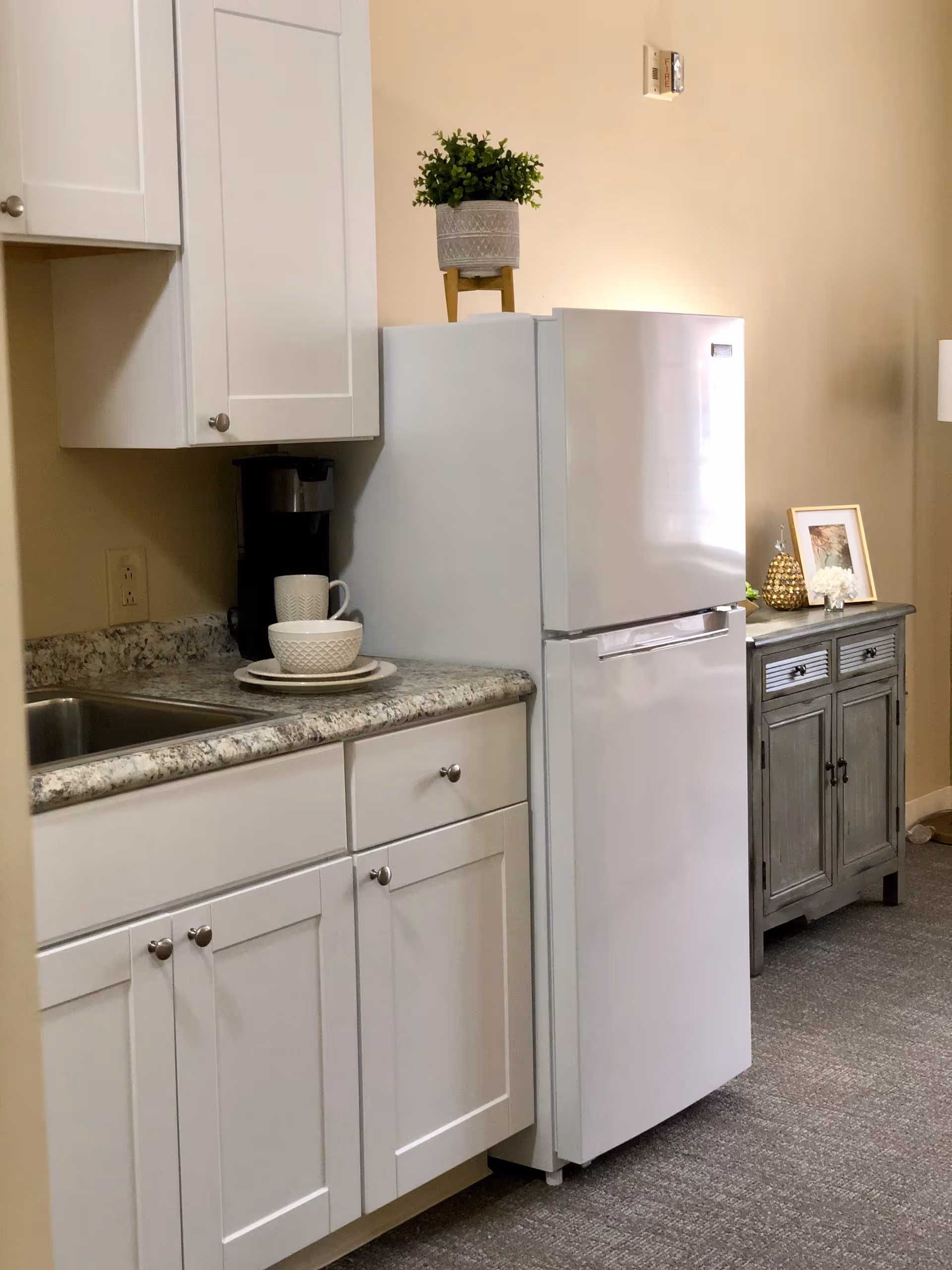A small kitchen area with white cabinets, a granite countertop, a stainless steel sink, a white refrigerator with a potted plant on top, and a coffee maker with a cup and bowl on the counter. In the background, there is a gray wooden sideboard with decorative items including a framed picture, a golden pineapple, and a small flower arrangement.